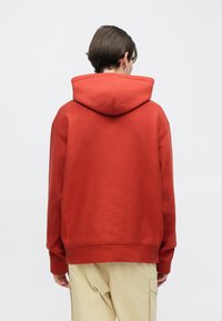 Κόκκινο φούτερ με μεγάλη κουκούλα, κατασκευασμένο από απαλό ύφασμα. Διαθέτει χαμηλωμένους ώμους και άνετη εφαρμογή, σε συνδυασμό με ανοιχτό μπεζ παντελόνι.