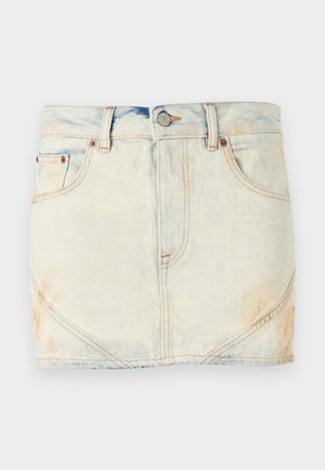 Henkilö, jolla on päällään vaalean beige nappipaita, vaalea farkku minihame ja mustat polven yli ulottuvat saappaat, seisoo yksinkertaisen valkoisen taustan edessä.