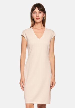 Frau trägt ein ärmelloses, knieumspielendes, hellbeiges Kleid mit V-Ausschnitt und goldenen Creolen, steht vor einem einfarbigen Hintergrund.