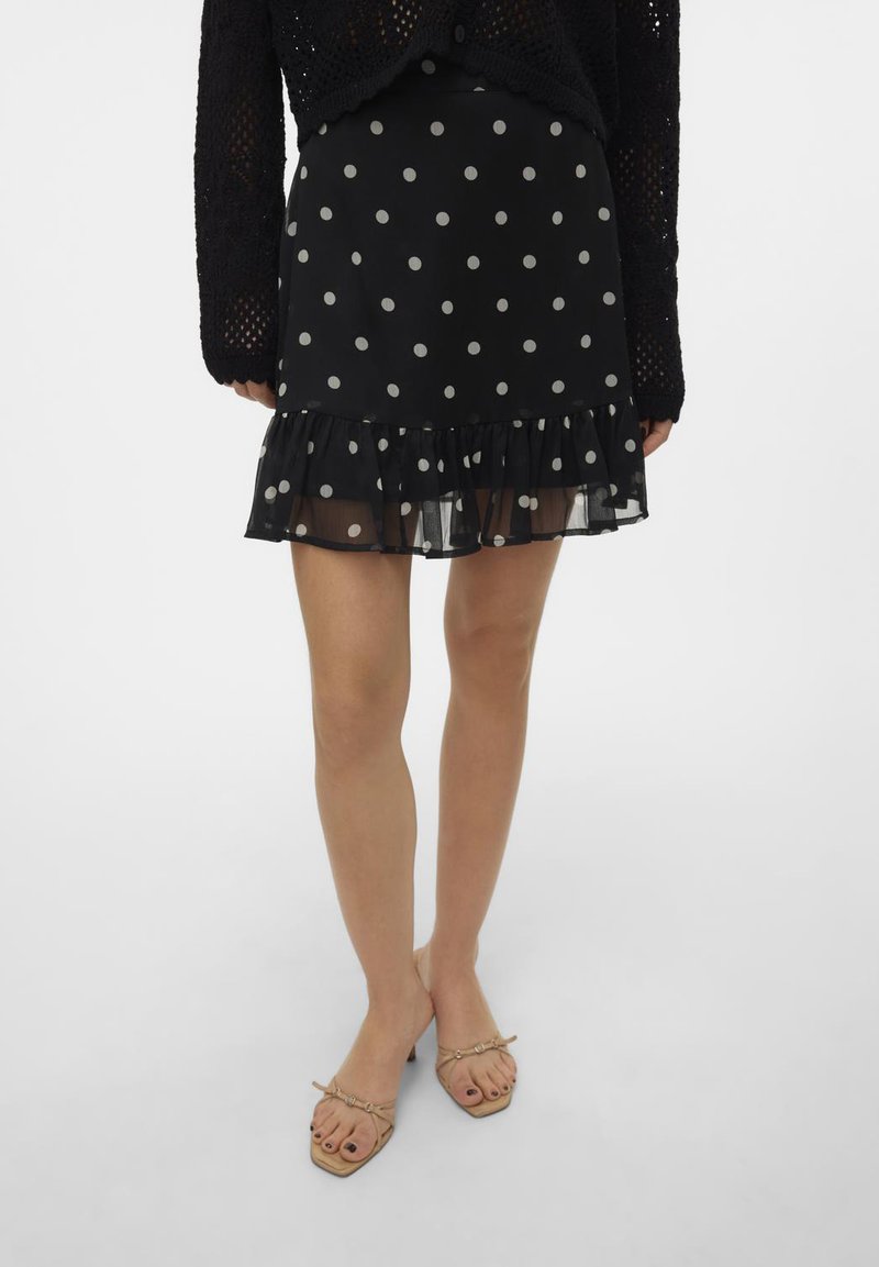 Zwarte rok met polka dots en een gerimpelde zoom, gemaakt van een doorschijnende stof. Gecombineerd met beige hakken. Witte achtergrond.