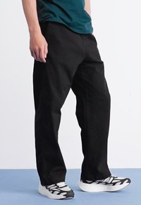 Personne portant un pantalon noir et des baskets noir et blanc, se tenant sur un sol bleu contre un fond blanc.