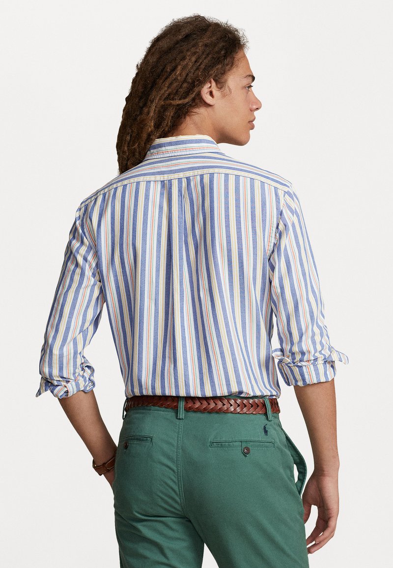 Gestreiftes Hemd mit blauen, gelben und weißen Streifen. Hochgeschlagene Ärmel, Rückenfalte, kombiniert mit grünen Hosen und einem geflochtenen braunen Gürtel.