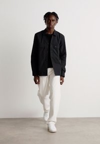 Chaqueta negra de botones con dos bolsillos en el pecho, llevada sobre una camisa negra. Combinada con pantalones blancos y zapatillas blancas sobre un fondo neutral.