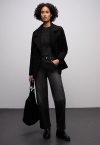 Černé sako s výstřihem s klopou, třpytivý šedý top, černé džíny s vysokým pasem a černé kotníkové boty. Drží černou tašku typu tote.