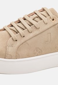 Beige skinnesko med preget mønster, med runde metalløyer, beige lisser og en hvit gummisåle for kontrast.