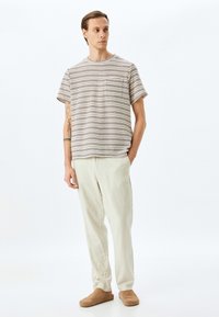 Camiseta de manga corta a rayas en beige y marrón oscuro, con un bolsillo en el pecho. Combinada con pantalones de lino beiges claros y zapatos marrones de tipo slip-on.