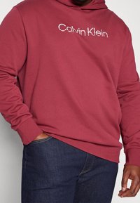 Bordowa bluza z kapturem wykonana z miękkiej tkaniny, z białym logo "Calvin Klein". Długie rękawy i luźny krój z kieszenią kangurką.