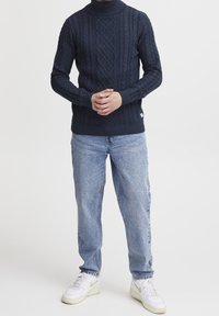 Personne portant un pull turtleneck en tricot torsadé bleu marine, un jean bleu clair et des baskets blanches avec un swoosh doré se tient les mains jointes.