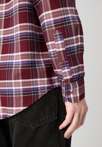 Camicia a scacchi nei colori bordeaux, blu e bianco. Presenta polsini con bottoni e orlo curvo. Indossata su un tessuto scuro, mette in evidenza un look a strati.