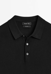 Polo shirt in cotone nero con colletto classico, dotato di tre bottoni argentati e un tessuto testurizzato. Etichetta del marchio visibile all'interno del colletto.