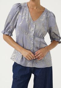 Blusa viola chiaro con motivo metallico oro, scollo a V, maniche a sbuffo e chiusura a bottoni. Abbinata a jeans a gamba larga di colore blu scuro.