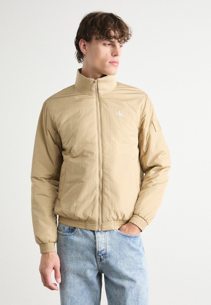 Giacca leggera beige con colletto alto, chiusura con zip e logo ricamato. Presenta polsini e vita elastici; tessuto liscio e strutturato.