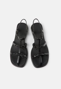 Black leather sandals with adjustable slingbacks, featuring flat soles and decorative bows on the straps. Smooth texture and simple design.