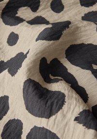 Tessuto beige con una texture opaca che presenta grandi macchie nere di leopardo in un motivo irregolare, creando un effetto visivo audace.