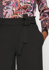 Zwarte hoog-gerande broek met een strikceintuur, met zijzakken en een gladde textuur. Gecombineerd met een blouse met een kleurrijk paisleypatroon.