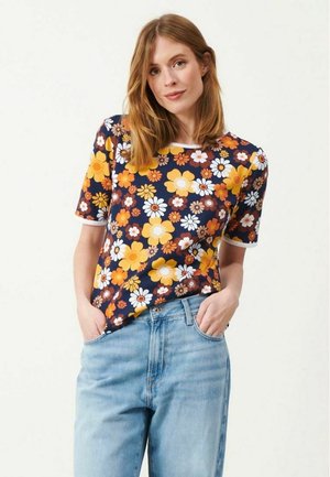 Kvinde med skulderlangt hår har en mørkeblå kortærmet skjorte med stort gult, hvidt og orange blomstermønster og lyseblå jeans med hænderne i lommerne på.