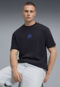 Jūras zilo kokvilnas T-kreklu ar īsām piedurknēm, kurā priekšpusē atrodas zils Marseļas logotips un Puma emblēma. Modelis valkā gaiši zilus džinsus.