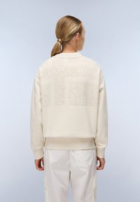 Crème-kleurige sweatshirt gemaakt van zachte stof met verhoogde patroon details op de achterkant, voorzien van een ronde halslijn en geribde mouwen.