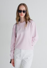 Light pink sweatshirt with round neckline and textured embroidery. Paired with white jeans and rectangular black sunglasses. Casual look.