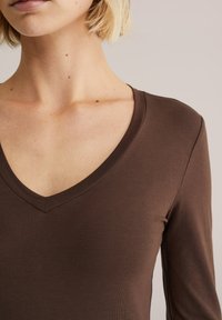 Barna hosszú ujjú felső V-nyakkivágással, sima, rugalmas anyagból készült. A nyakvonal és az ujjak végénél tisztán befejezett varrás található.