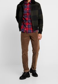 Chaqueta bomber de cuadros negros y verdes con un cuello suave sobre una camisa de cuadros rojos y azules, combinada con pantalones de pana marrones y zapatos negros.