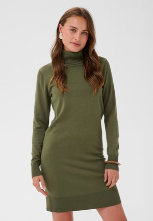 Femme aux longs cheveux bruns portant une robe pull à col roulé vert olive, des boucles d'oreilles blanches et un bracelet en bois, debout devant un fond uni.