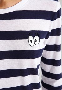 Pasiasty longsleeve w kolorze granatowym i białym z poziomymi paskami, okrągłym dekoltem oraz małym haftowanym rysunkiem oka na klatce piersiowej.