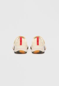 Beige sneakers med teksturert overdel og rød pull-tab, med en kraftig såle med en gjennomsiktig del og subtile mønsterdetaljer.