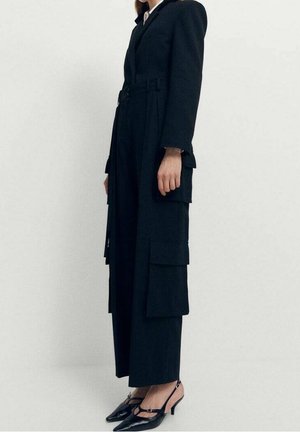 Kobieta ubrana w długi czarny płaszcz z dużymi kieszeniami i czarne, spiczaste sandały na obcasie, stojąca na tle jednolitego białego tła.