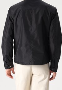 Černá lehká bunda s hladkým povrchem, se standardním límcem a manžetami na knoflíky, zobrazená zezadu. Minimalistický design.