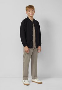 Chłopiec stoi w oparciu o szary mur, ubrany w czarną kurtkę na guziki, beżowe spodnie, beżową koszulę i białe trampki z gumową podeszwą.