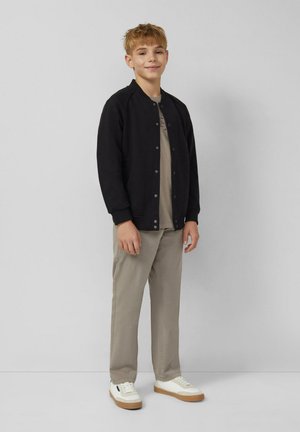 Chłopiec stoi w oparciu o szary mur, ubrany w czarną kurtkę na guziki, beżowe spodnie, beżową koszulę i białe trampki z gumową podeszwą.