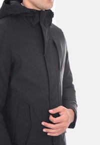 Giacca nera impermeabile con colletto alto, cappuccio regolabile e tasche laterali. Presenta una texture liscia e un design minimalista.