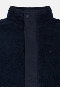 Sötétkék bolyhos dzseki magas gallérral, texturált külső anyaggal és sima belső béléssel, elöl gombos záródással.