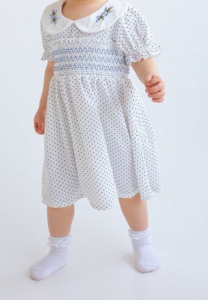 Tout-petit portant une robe blanche à pois noirs, un col brodé et des chaussettes blanches avec bordure en dentelle, debout sur un fond uni.