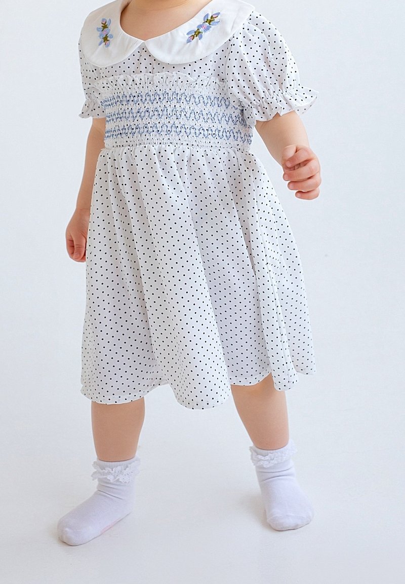 Tout-petit portant une robe blanche à pois noirs, un col brodé et des chaussettes blanches avec bordure en dentelle, debout sur un fond uni.