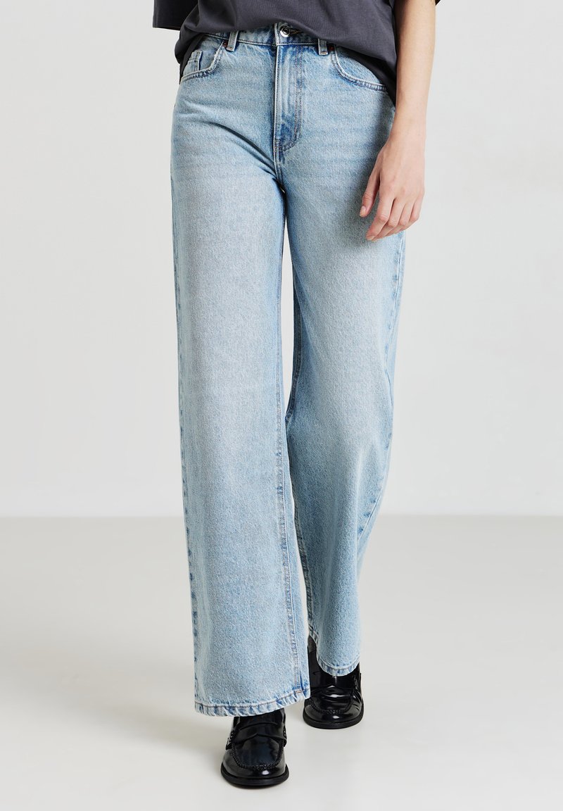 Jean large bleu clair en denim, avec une taille haute, design classique à cinq poches et ourlet effiloché. Portés avec des chaussures noires.