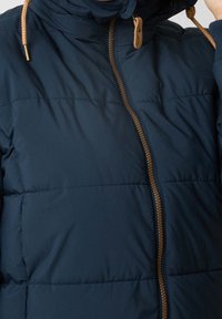 Veste matelassée marine avec une finition texturée, dotée d'une fermeture éclair à l'avant avec des accents beiges et d'une capuche à cordon de serrage. Isolée pour la chaleur.