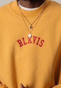 Sudadera amarilla con el texto bordado en rojo "BLKVIS" en forma de arco en la parte delantera. Presenta un cuello redondo y capas de collares de oro.