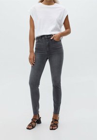 Frau trägt ein weißes ärmelloses Oberteil, hochgeschnittene, ausgewaschene schwarze Skinny-Jeans und schwarze, geschlossene Blockabsatzsandalen, steht vor einem schlichten Hintergrund.