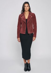 Vrouw staand, gekleed in een bordeauxrode suède bikerjack, zwarte midi-jurk en zwarte enkellaarsjes tegen een eenvoudige lichte achtergrond.