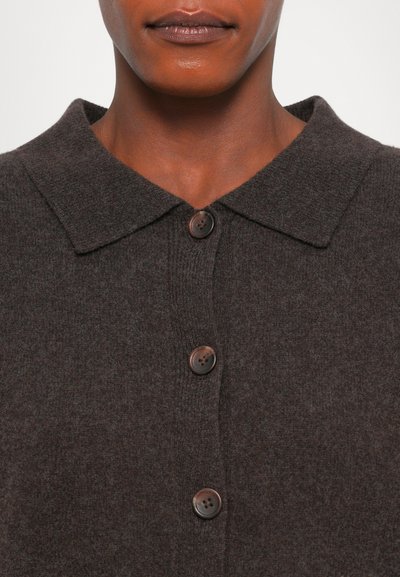 Gros plan sur une personne portant un cardigan tricoté à boutons de couleur marron foncé avec un large col et trois boutons visibles.