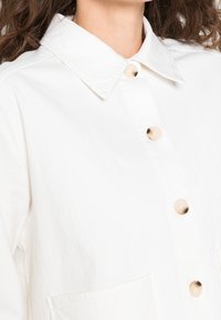 Camisa blanca de botones con cuello, que presenta grandes botones color crema y una tela texturizada. Dos bolsillos frontales visibles.