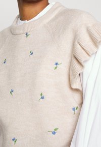 Beżowy sweter z dzianiny z niebieskim haftem kwiatowym, okrągłym dekoltem i fakturowanymi falbanami na rękawach.