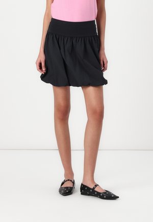 Woman wearing a black gathered mini skirt, pink top, and black pointed flats with silver eyelet details, standing against a white background.