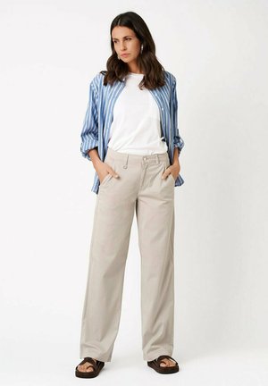 Kobieta stoi z rękami w kieszeniach, ubrana w białą koszulkę, niebiesko-białą pasekowaną otwartą koszulę, beżowe spodnie z szerokimi nogawkami i brązowe sandały.