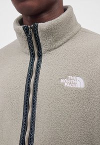 Gros plan d'une personne portant une veste polaire beige avec une fermeture éclair noire et le logo blanc The North Face sur la poitrine.