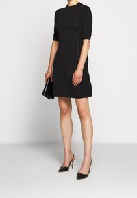 Femme en robe noire longueur genoux et escarpins noirs tenant une pochette noire, debout devant un fond blanc uni.