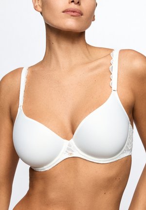 Vrouw die een witte beha met gladde cups en geschulpte randen op de bandjes draagt, waarbij sleutelbeenderen en bovenlichaam zichtbaar zijn tegen een witte achtergrond.