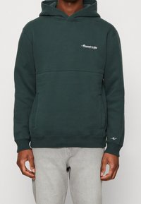 Haut à capuche vert foncé en tissu en mélange de coton avec une poche avant, des poignets côtelés et un petit logo sur la poitrine. Texture lisse.